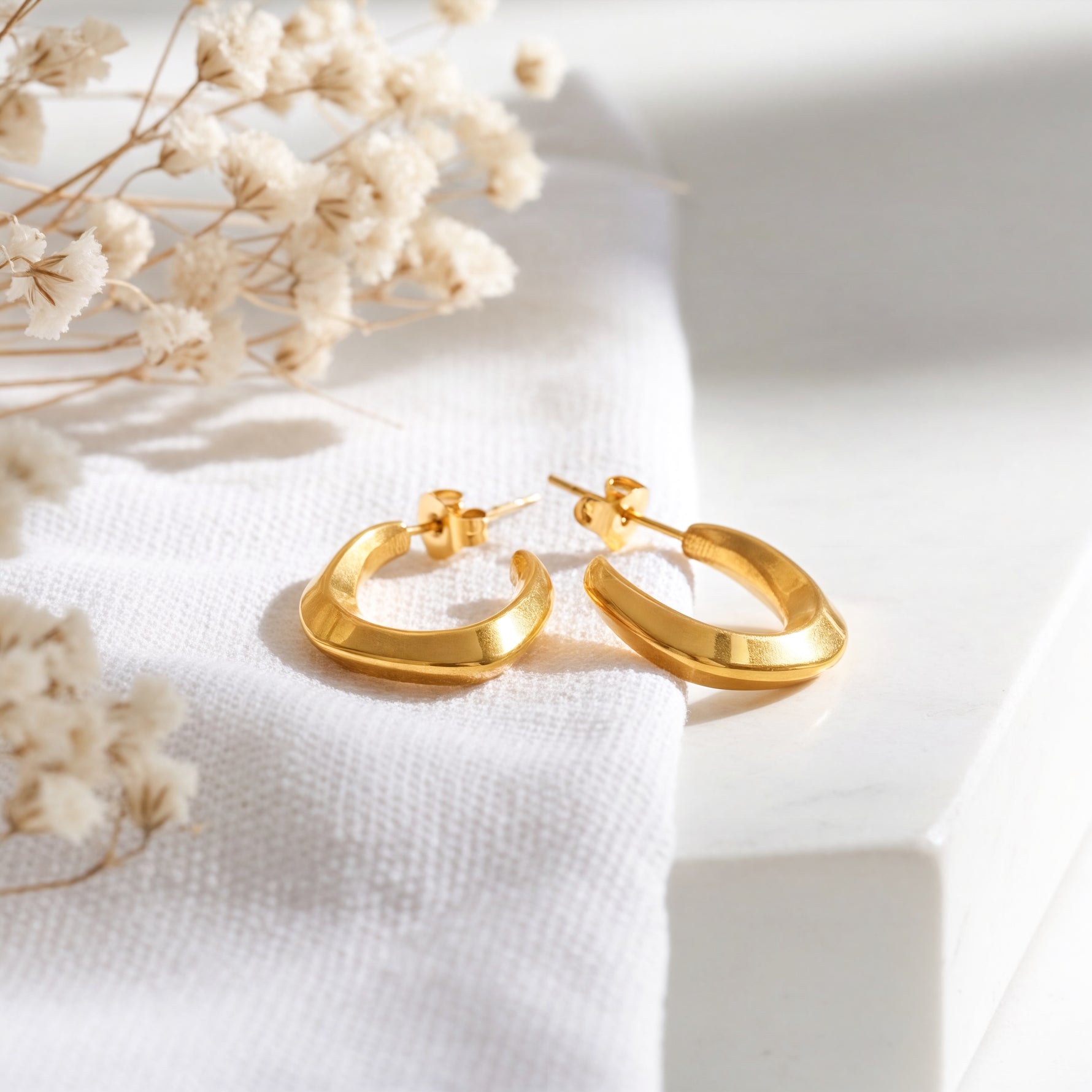 Geometrische Creolen mit kantigem Design aus Edelstahl 18 K vergoldet, liegen auf hellem Stoff mit Trockenblumen im Hintergrund – Virel Creolen | Mercy’s Jewelry