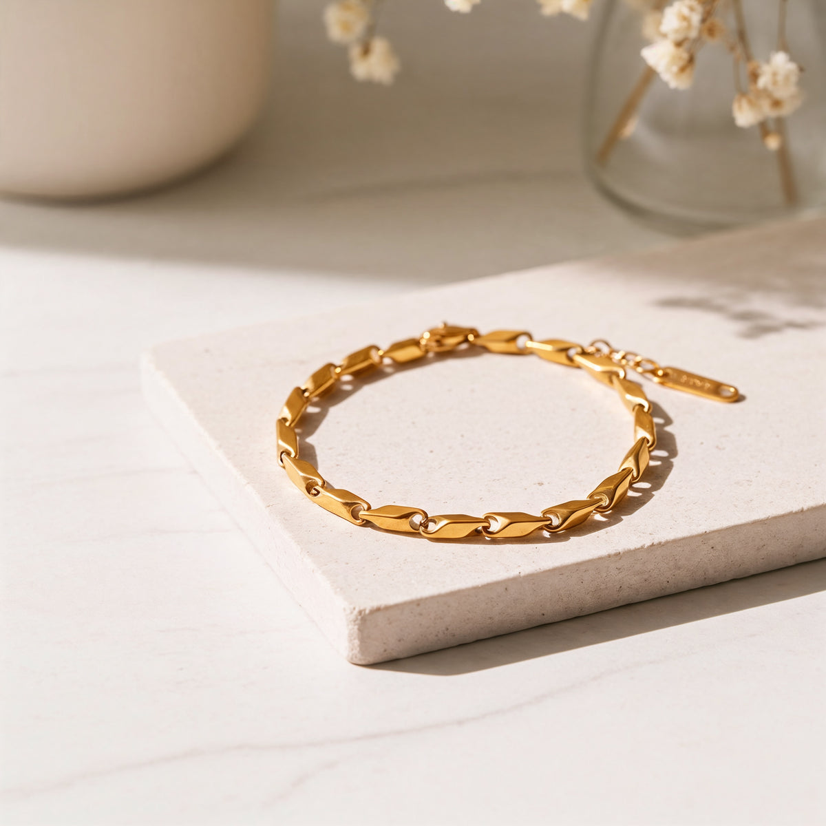 Gliederarmband aus Edelstahl 18 K vergoldet mit flachen, modernen Gliedern, liegend auf heller Steinplatte mit Trockenblumen im Hintergrund – Talea Armband | Mercy’s Jewelry