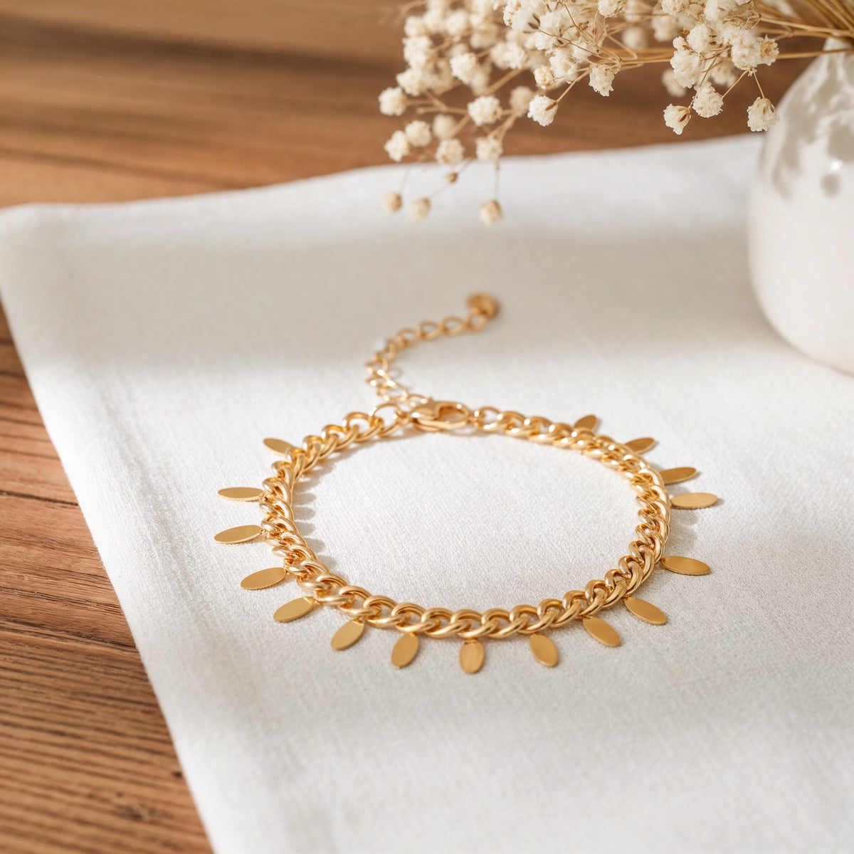 Gliederarmband aus Edelstahl 18 K vergoldet mit flachen Plättchen-Anhängerchen, liegend auf weißem Stoff mit Trockenblumen im Hintergrund – Marvella Armband | Mercy’s Jewelry
