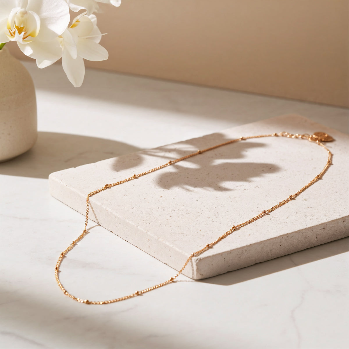 Feine Gliederhalskette aus Edelstahl 18 K vergoldet, liegt auf heller Steinplatte mit weißen Blumen im Hintergrund – Enira Halskette | Mercy’s Jewelry