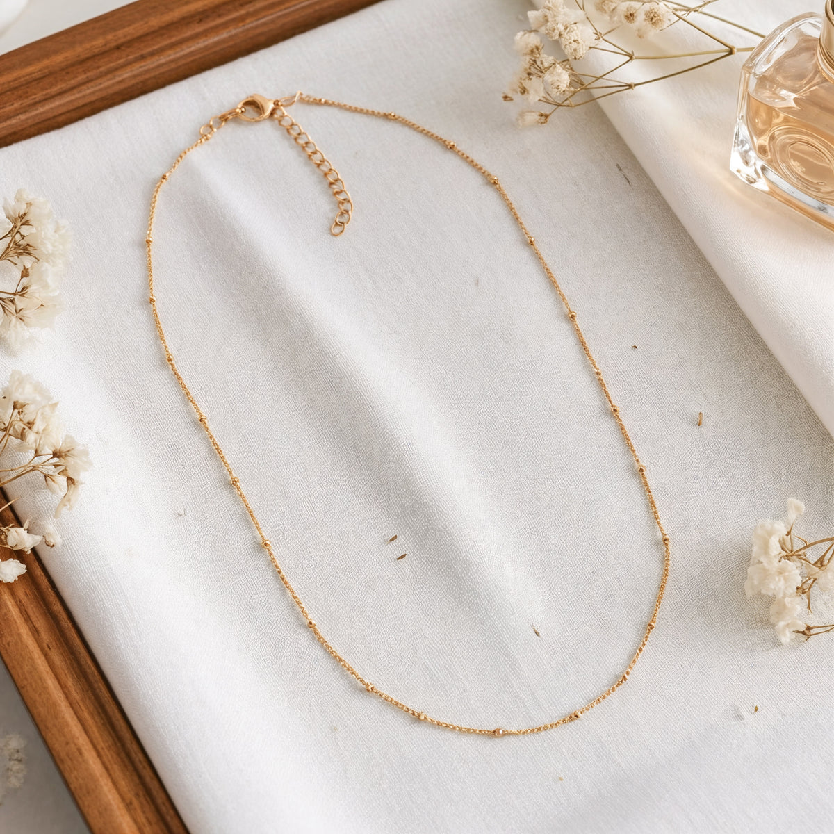 Feine Gliederhalskette aus Edelstahl 18 K vergoldet, liegt auf weißem Stoff mit Holzrahmen, Trockenblumen und Parfümflakon – Enira Halskette | Mercy’s Jewelry