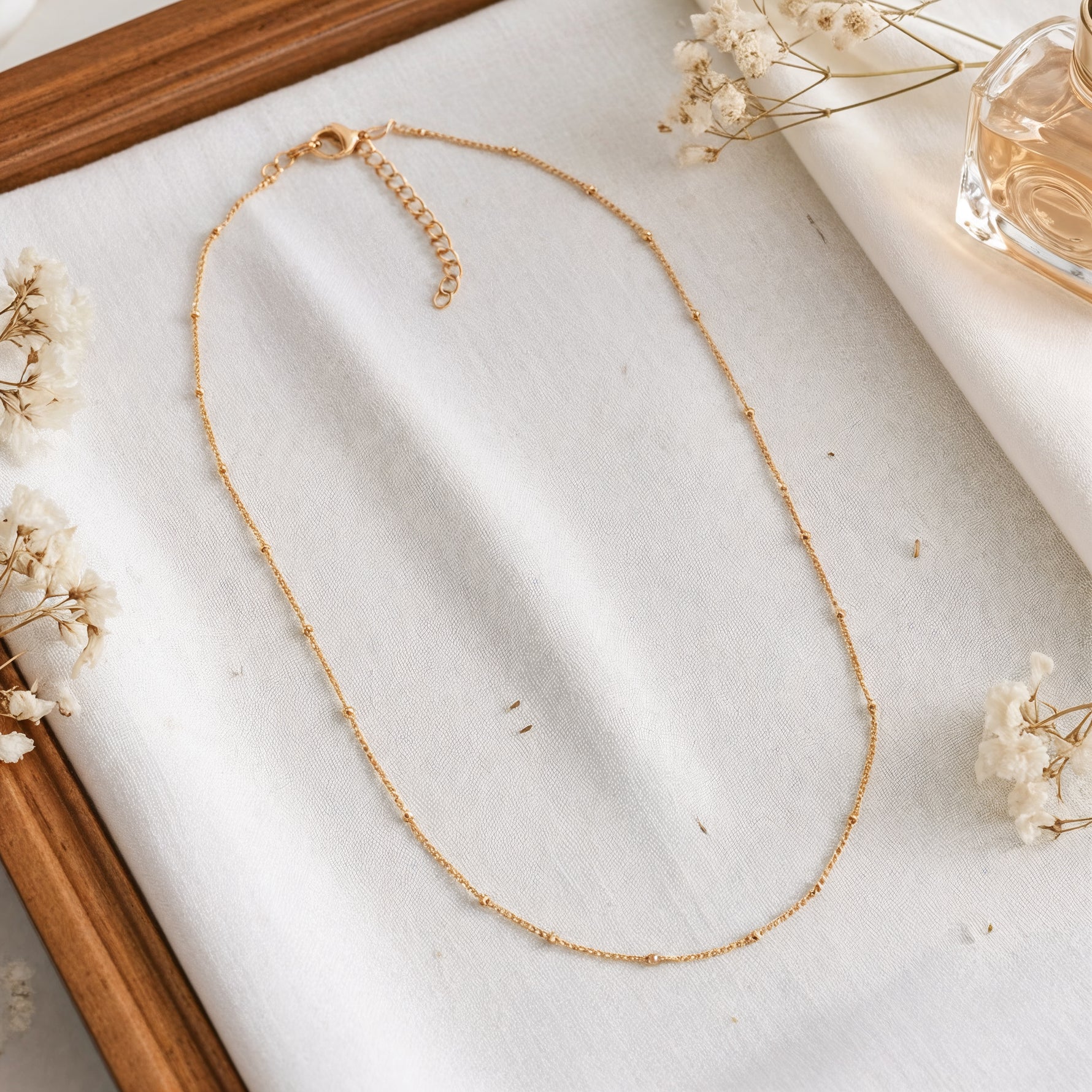 Feine Gliederhalskette aus Edelstahl 18 K vergoldet, liegt auf weißem Stoff mit Holzrahmen, Trockenblumen und Parfümflakon – Enira Halskette | Mercy’s Jewelry