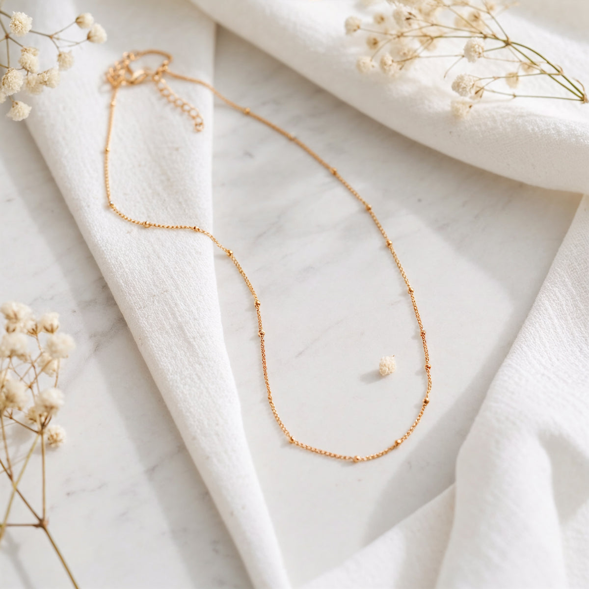 Feine Gliederhalskette aus Edelstahl 18 K vergoldet, liegt auf weißem Stoff mit Marmorhintergrund und getrockneten Blumen – Enira Halskette | Mercy’s Jewelry