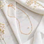 Feine Gliederhalskette aus Edelstahl 18 K vergoldet, liegt auf weißem Stoff mit Marmorhintergrund und getrockneten Blumen – Enira Halskette | Mercy’s Jewelry