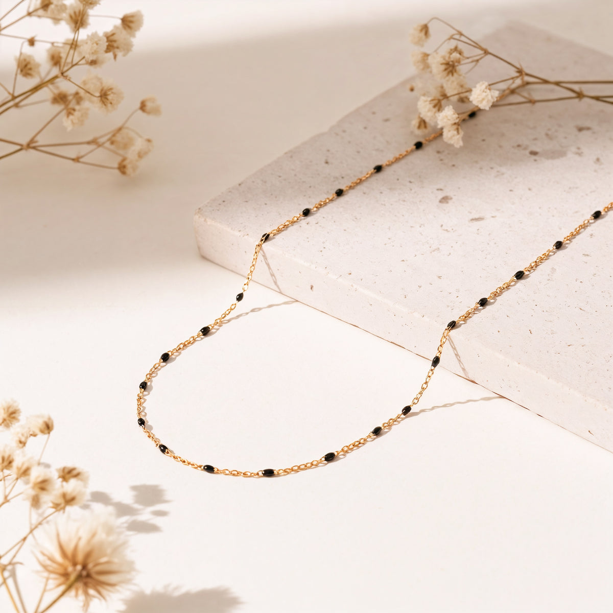 Feine Gliederhalskette mit schwarzen Perlen aus Edelstahl 18 K vergoldet, liegt auf heller Steinplatte mit getrockneten Blumen – Nivra Halskette | Mercy’s Jewelry