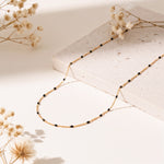 Feine Gliederhalskette mit schwarzen Perlen aus Edelstahl 18 K vergoldet, liegt auf heller Steinplatte mit getrockneten Blumen – Nivra Halskette | Mercy’s Jewelry