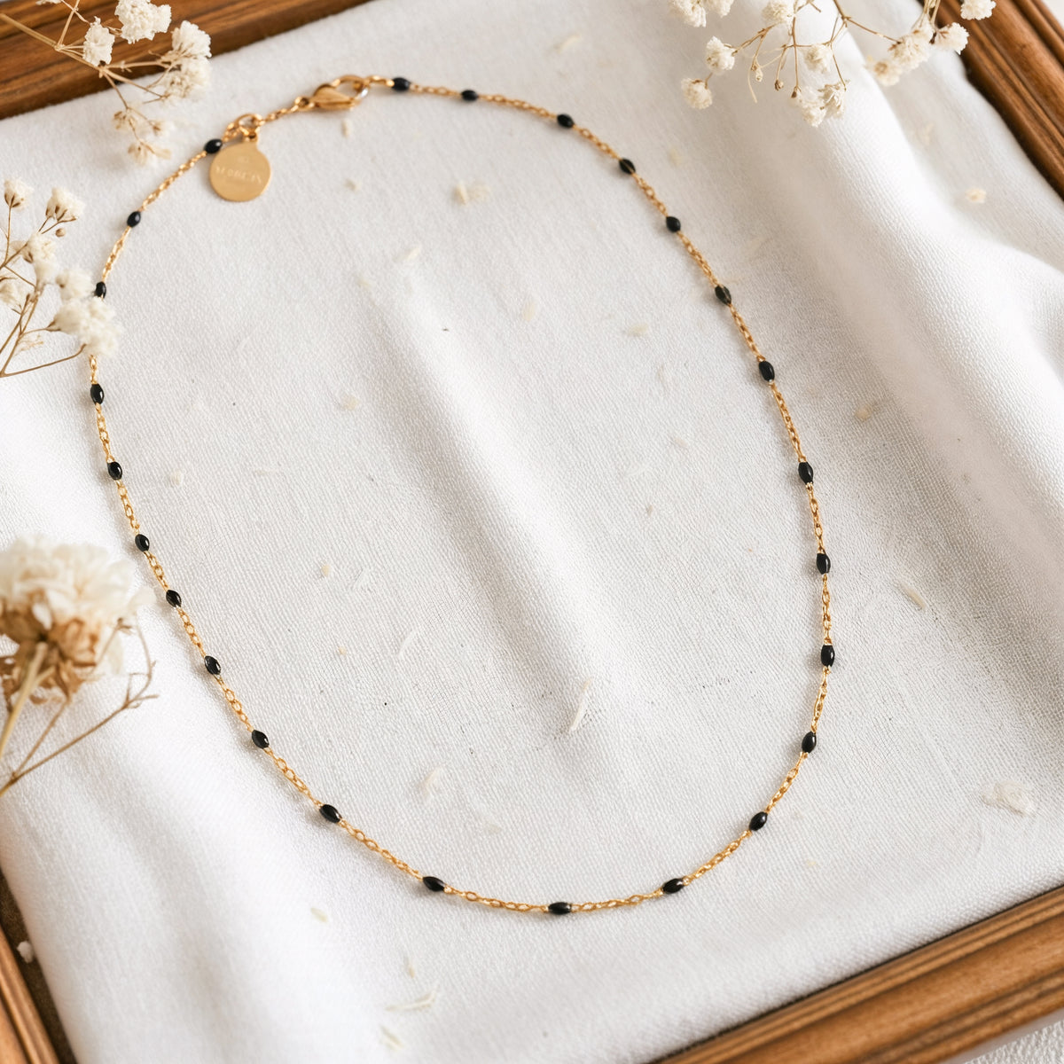Feine Gliederhalskette mit schwarzen Perlen aus Edelstahl 18 K vergoldet, liegt auf weißem Stoff mit Holzhintergrund und getrockneten Blumen – Nivra Halskette | Mercy’s Jewelry