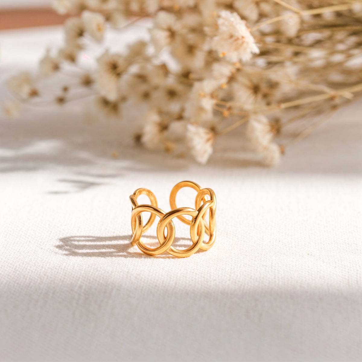 Gliederring aus Edelstahl 18 K vergoldet, mit ineinander verschlungenem Design auf heller Stoffoberfläche und Trockenblumen im Hintergrund – Eloura Ring | Mercy’s Jewelry