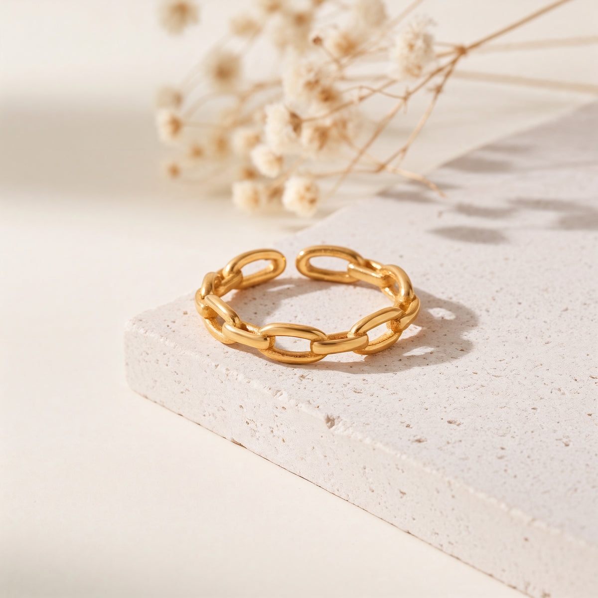 Gliederring aus Edelstahl 18 K vergoldet, offen und verstellbar, mit strukturierter Kettenoptik auf heller Steinplattform mit Trockenblumen im Hintergrund – Olyra Ring | Mercy’s Jewelry