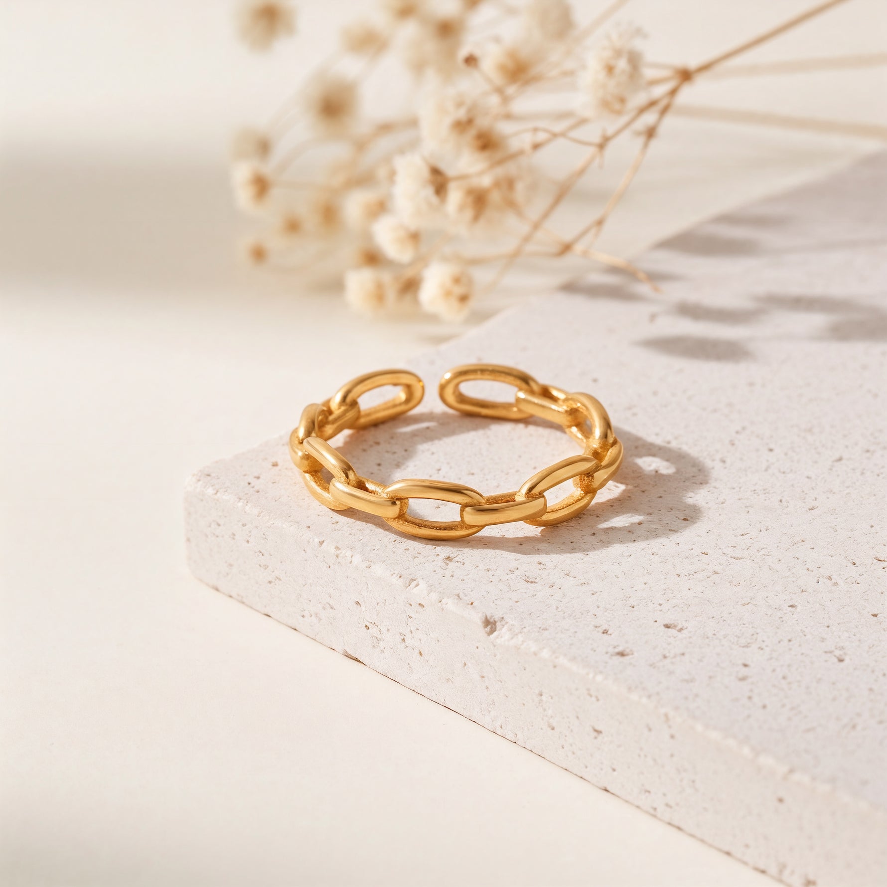 Gliederring aus Edelstahl 18 K vergoldet, offen und verstellbar, mit strukturierter Kettenoptik auf heller Steinplattform mit Trockenblumen im Hintergrund – Olyra Ring | Mercy’s Jewelry
