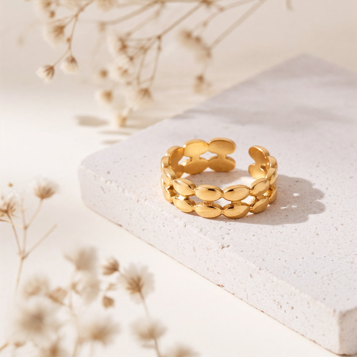 Gliederring aus Edelstahl 18 K vergoldet, offen und verstellbar, mit ovalen Glied-Elementen auf heller Steinplattform mit Trockenblumen im Hintergrund – Seraya Ring | Mercy’s Jewelry