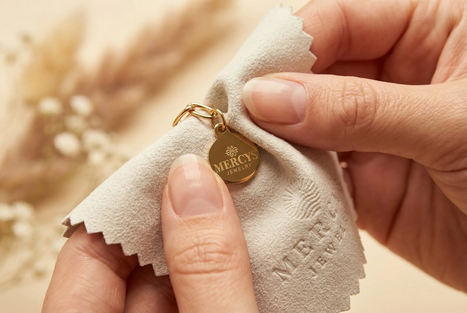 Hände polieren einen goldenen Mercy’s Jewelry Anhänger mit einem weichen Poliertuch, umgeben von Pampasgras und Baby’s Breath im warmen Licht.