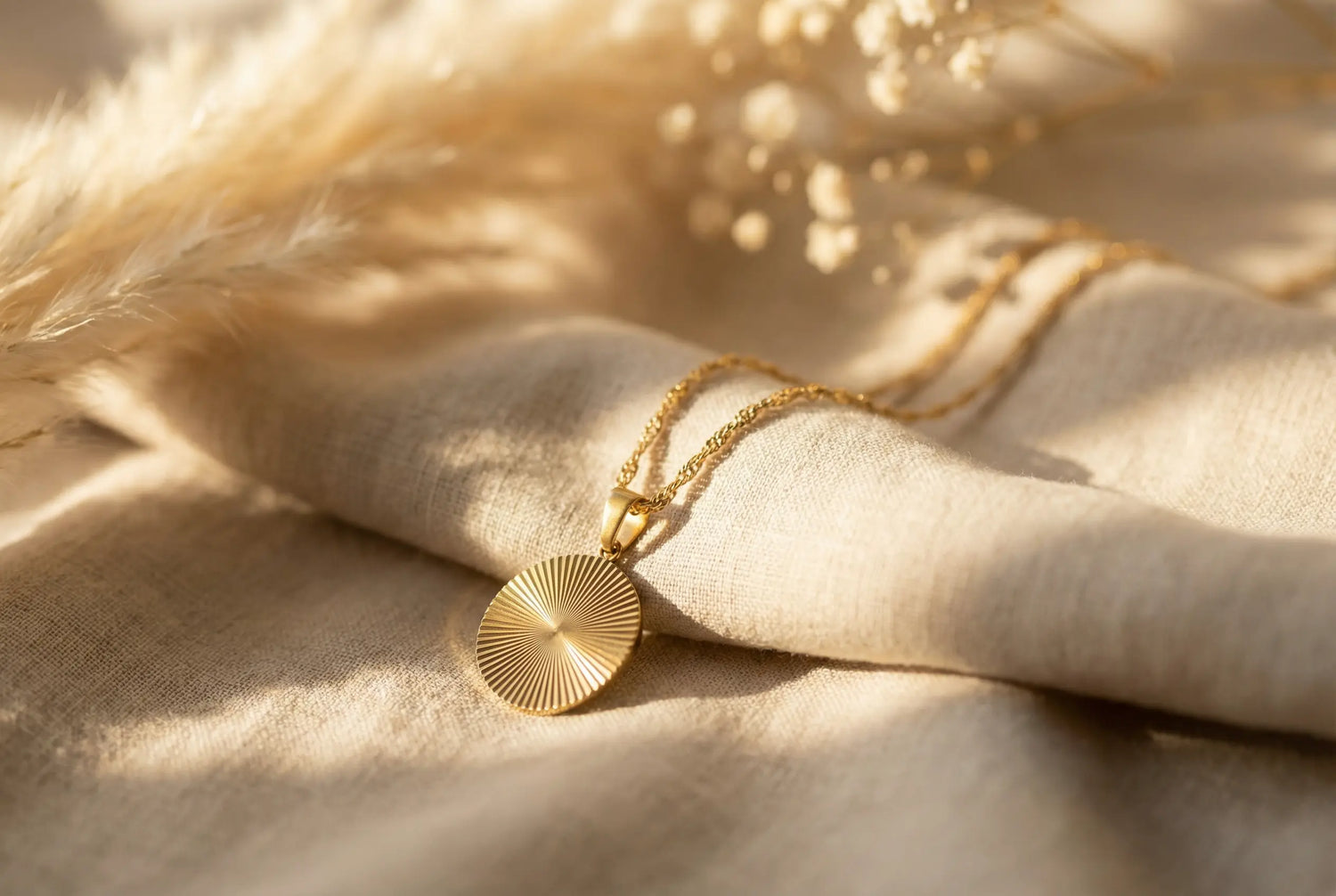 Goldene Kette mit rundem Sonnenstrahl-Anhänger auf weichem Leinenstoff, dekoriert mit Pampasgras und Baby’s Breath im warmen Licht.