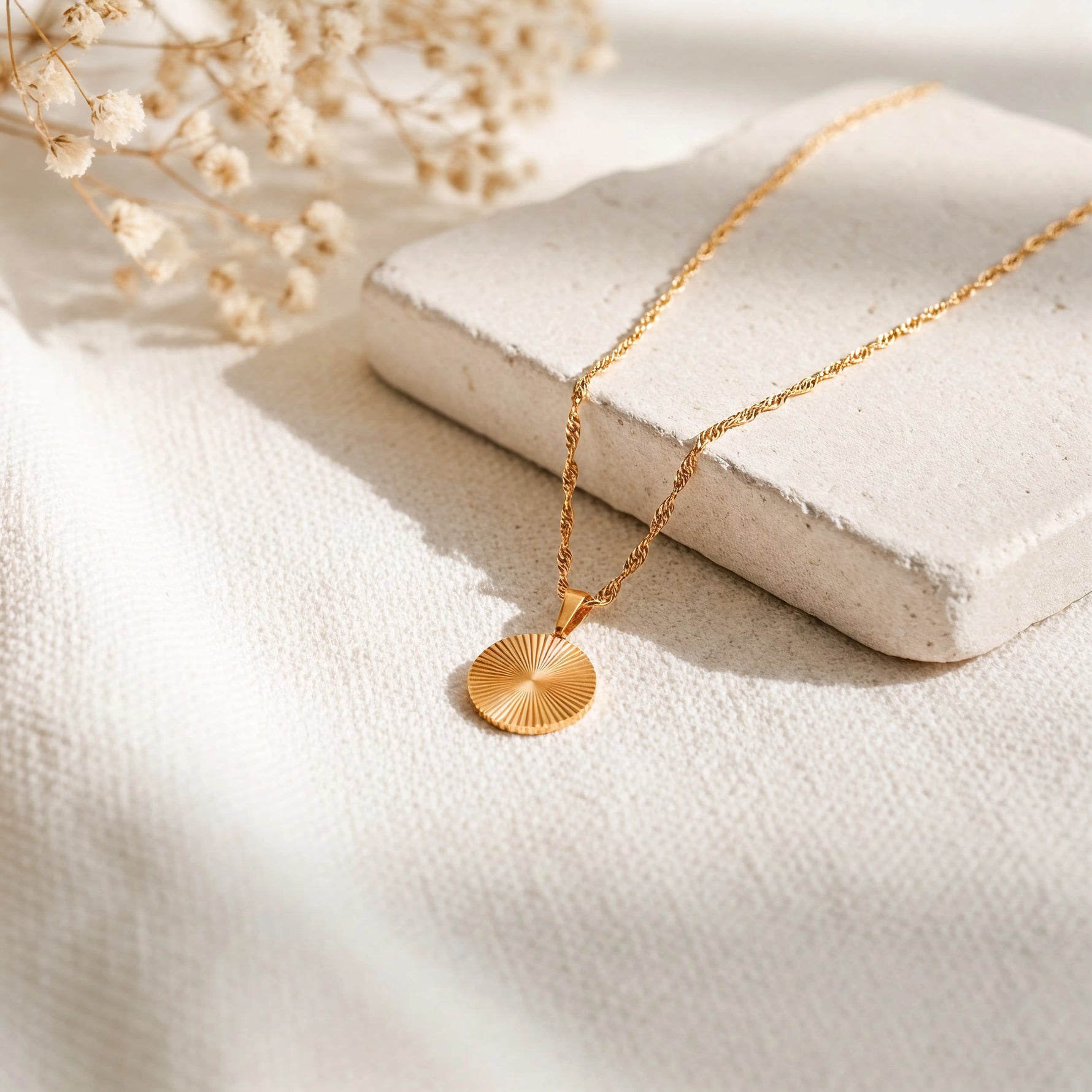 Goldene Halskette mit rundem Sonnenstrahl-Anhänger auf einem hellen Steinpodest, umgeben von Leinenstoff und Baby’s Breath im weichen Tageslicht.