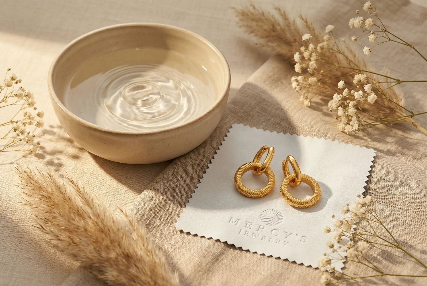 Goldene Ohrringe im Ringdesign auf einem Mercy’s Jewelry Poliertuch neben einer Keramikschale mit Wasser, umgeben von Pampasgras und Baby’s Breath im warmen Licht.