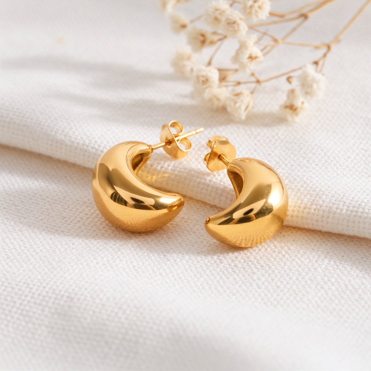 Halbcreolen mit rundem, voluminösem Design aus Edelstahl 18 K vergoldet, liegen auf hellem Stoff mit Trockenblumen im Hintergrund – Nayen Halbcreolen | Mercy’s Jewelry