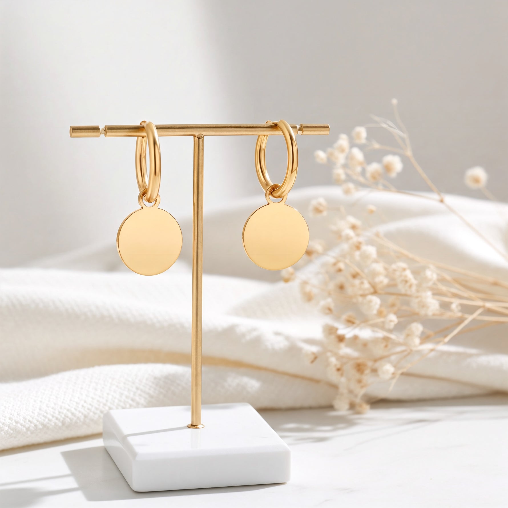 Huggie-Creolen aus Edelstahl 18 K vergoldet mit runden Plättchen-Anhängern, hängen an goldenem Ständer auf hellem Stoff mit Trockenblumen im Hintergrund – Isara Huggies | Mercy’s Jewelry