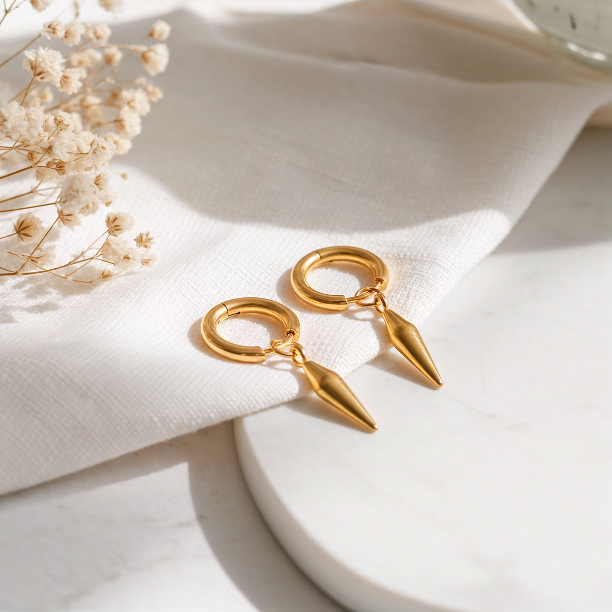 Huggie-Ohrringe mit abnehmbaren Spike-Anhänger aus Edelstahl 18 K vergoldet, liegen auf hellem Leinentuch mit Trockenblumen im Sonnenlicht – Nyros Huggies | Mercy’s Jewelry