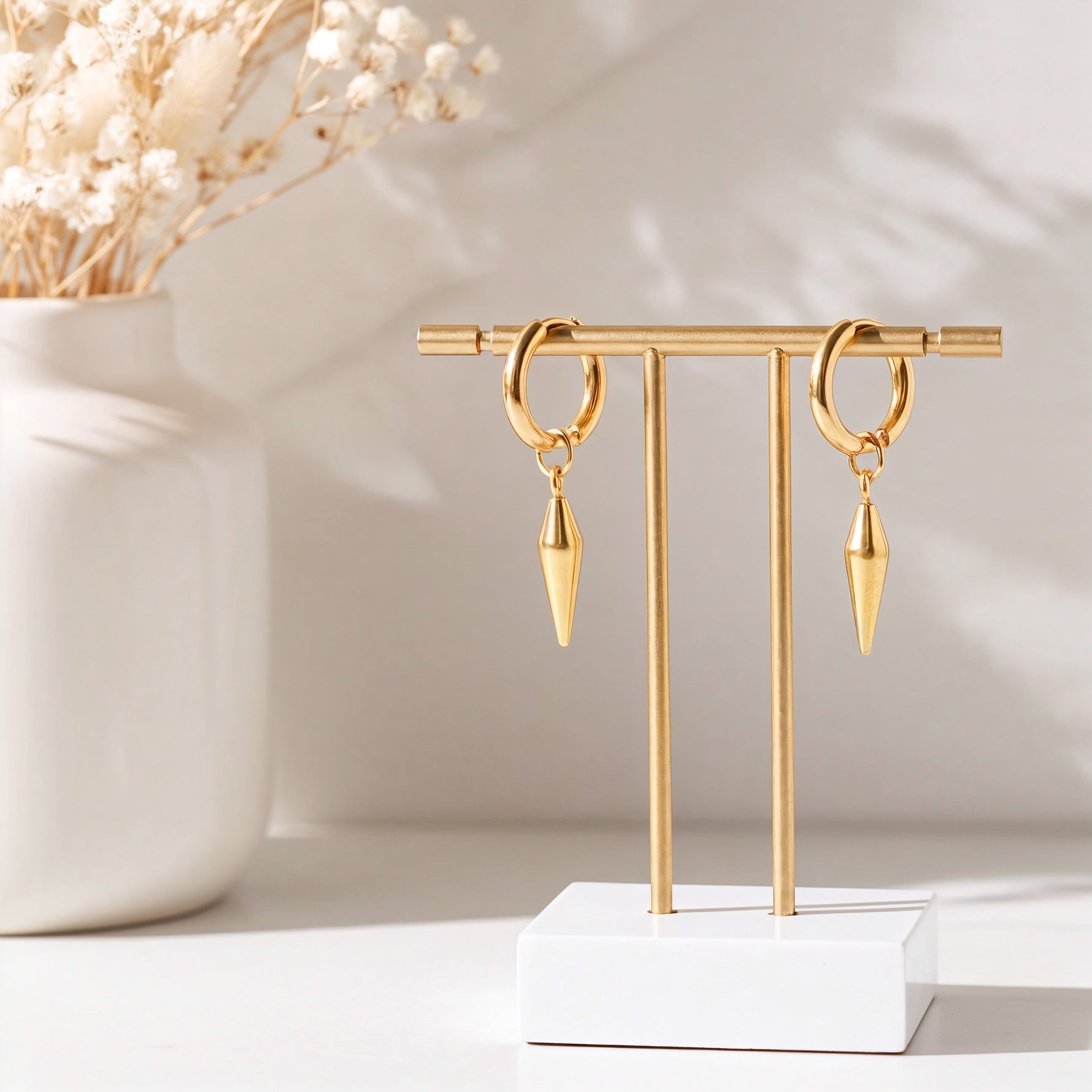 Huggie-Ohrringe mit abnehmbaren Spike-Anhänger aus Edelstahl 18 K vergoldet, hängen an goldfarbenem Ständer mit hellem Hintergrund und Trockenblumen – Nyros Huggies | Mercy’s Jewelry