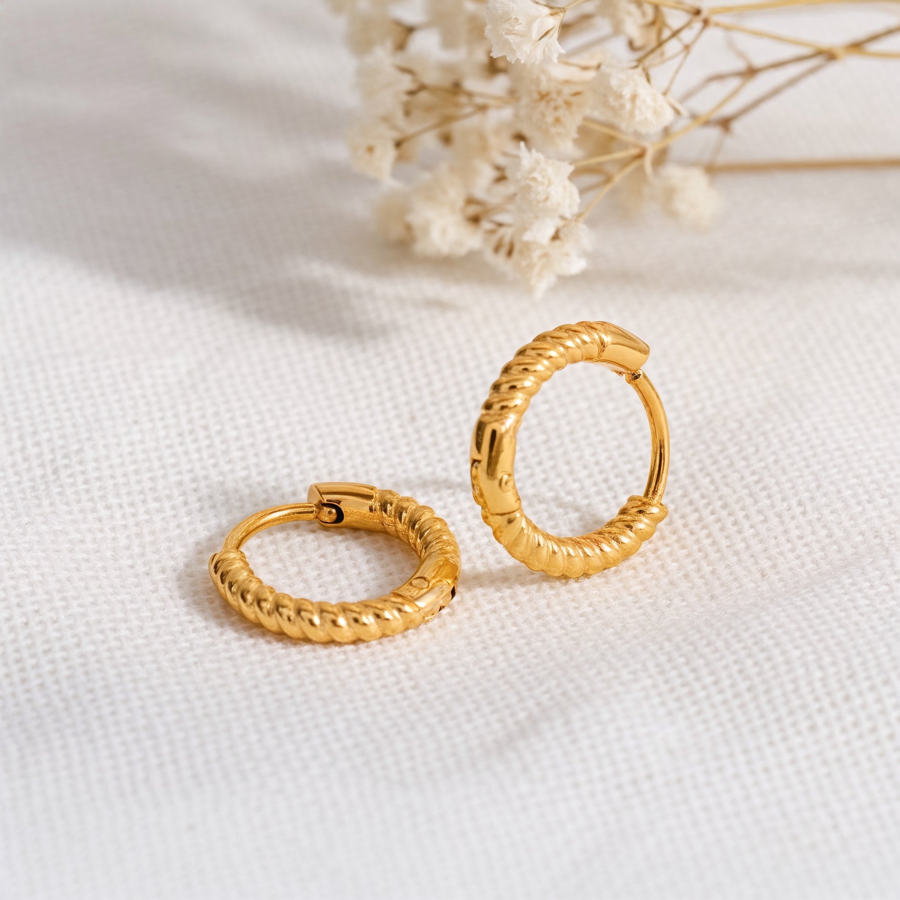 Strukturierte Huggie-Creolen mit seilartigem Design aus Edelstahl 18 K vergoldet, liegen auf hellem Stoff mit Trockenblumen im Hintergrund – Avenil Huggies | Mercy’s Jewelry