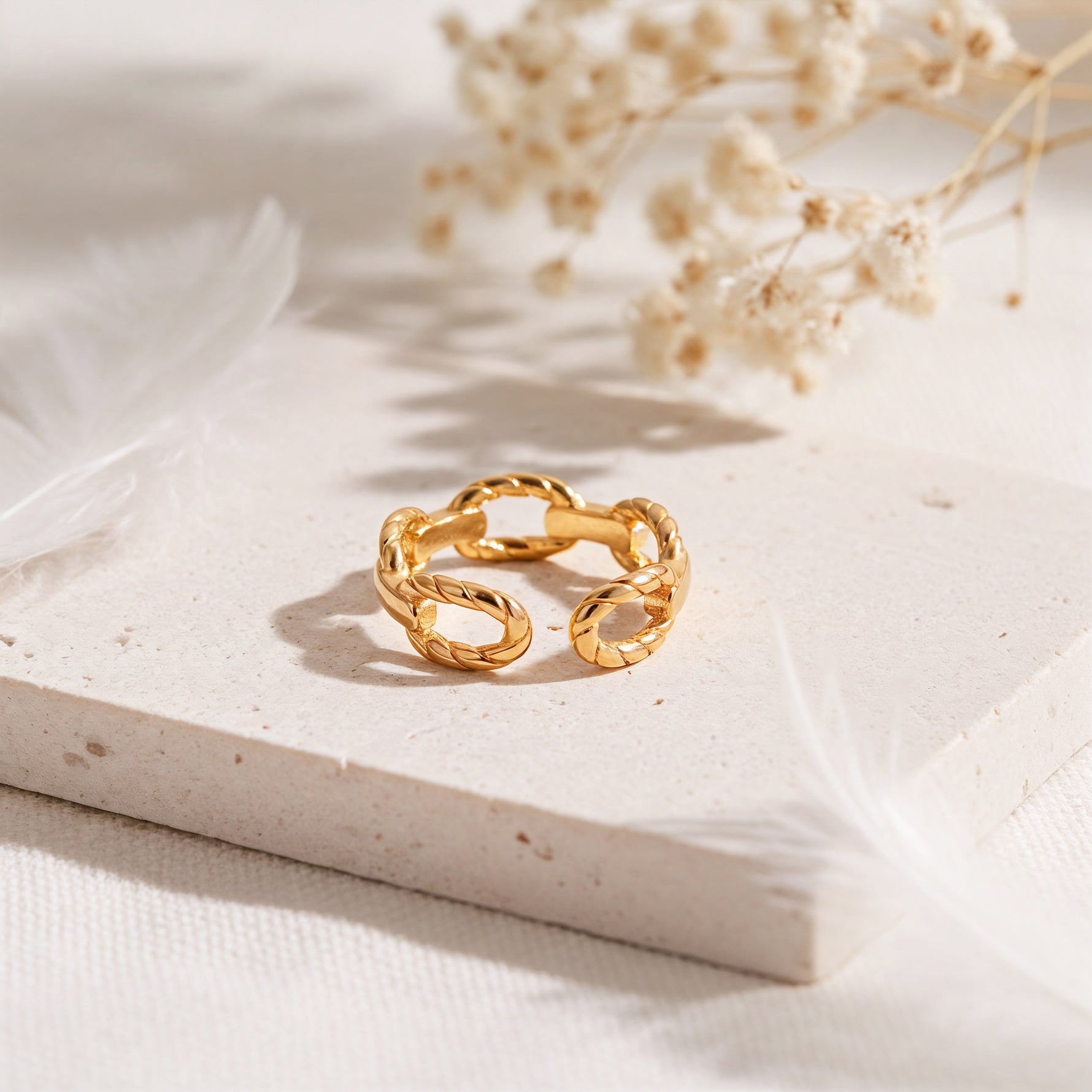 Kettenring aus Edelstahl 18 K vergoldet, offen und verstellbar, mit gedrehten Gliedern im modernen Design auf heller Steinplattform mit Trockenblumen und Feder im Vordergrund – Talira Ring | Mercy’s Jewelry