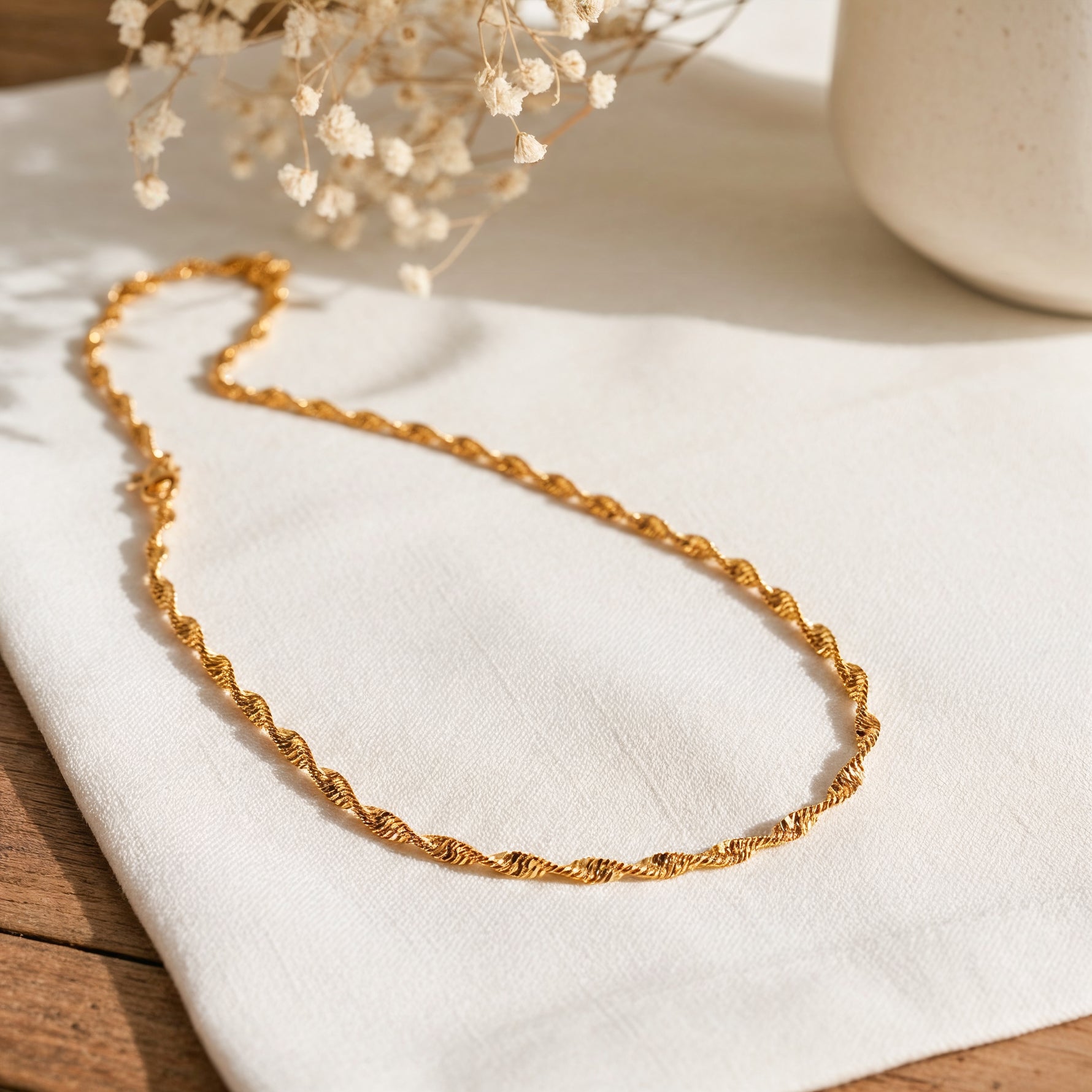 Kordel-Halskette aus Edelstahl 18 K vergoldet, liegt auf hellem Stoff und Holztisch mit Trockenblumen und Vase im Hintergrund – Solira Halskette | Mercy’s Jewelry