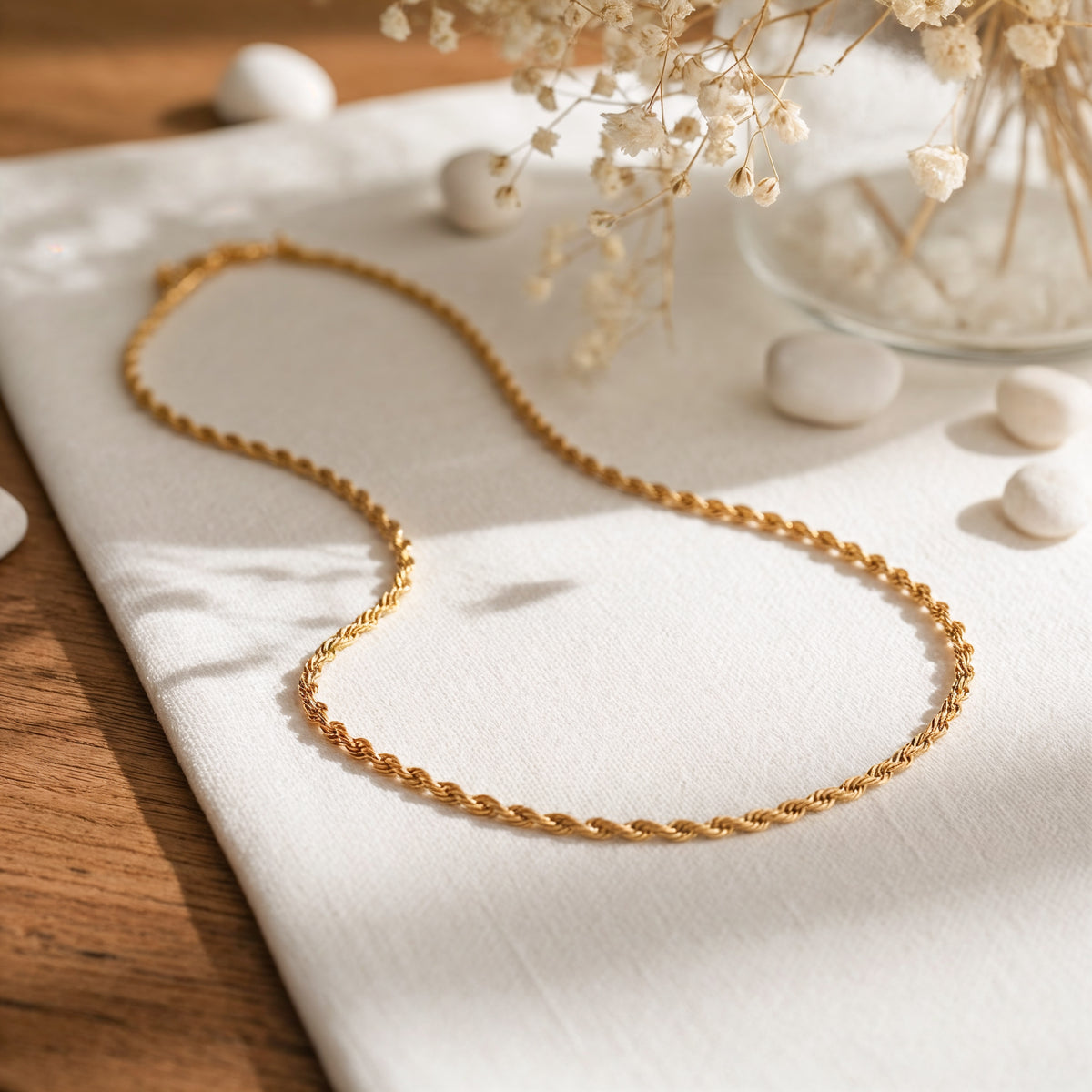 Goldene Kordel-Halskette aus Edelstahl 18 K vergoldet, liegt auf weißem Stoff mit Holzhintergrund und getrockneten Blumen – Tharien Halskette | Mercy’s Jewelry