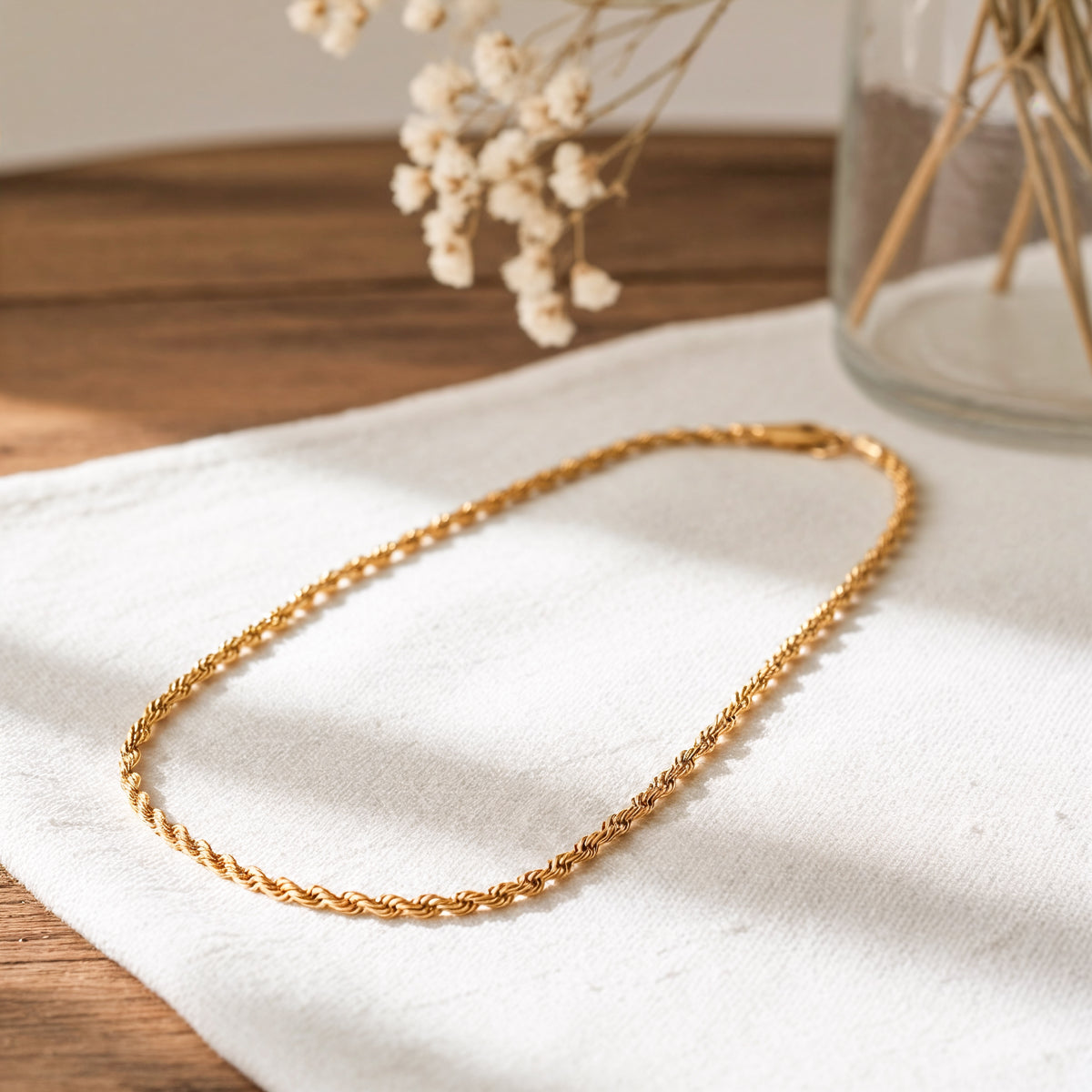 Goldene Kordel-Halskette aus Edelstahl 18 K vergoldet, liegt auf weißem Stoff mit Holzhintergrund und Trockenblumen im Glas – Tharien Halskette | Mercy’s Jewelry
