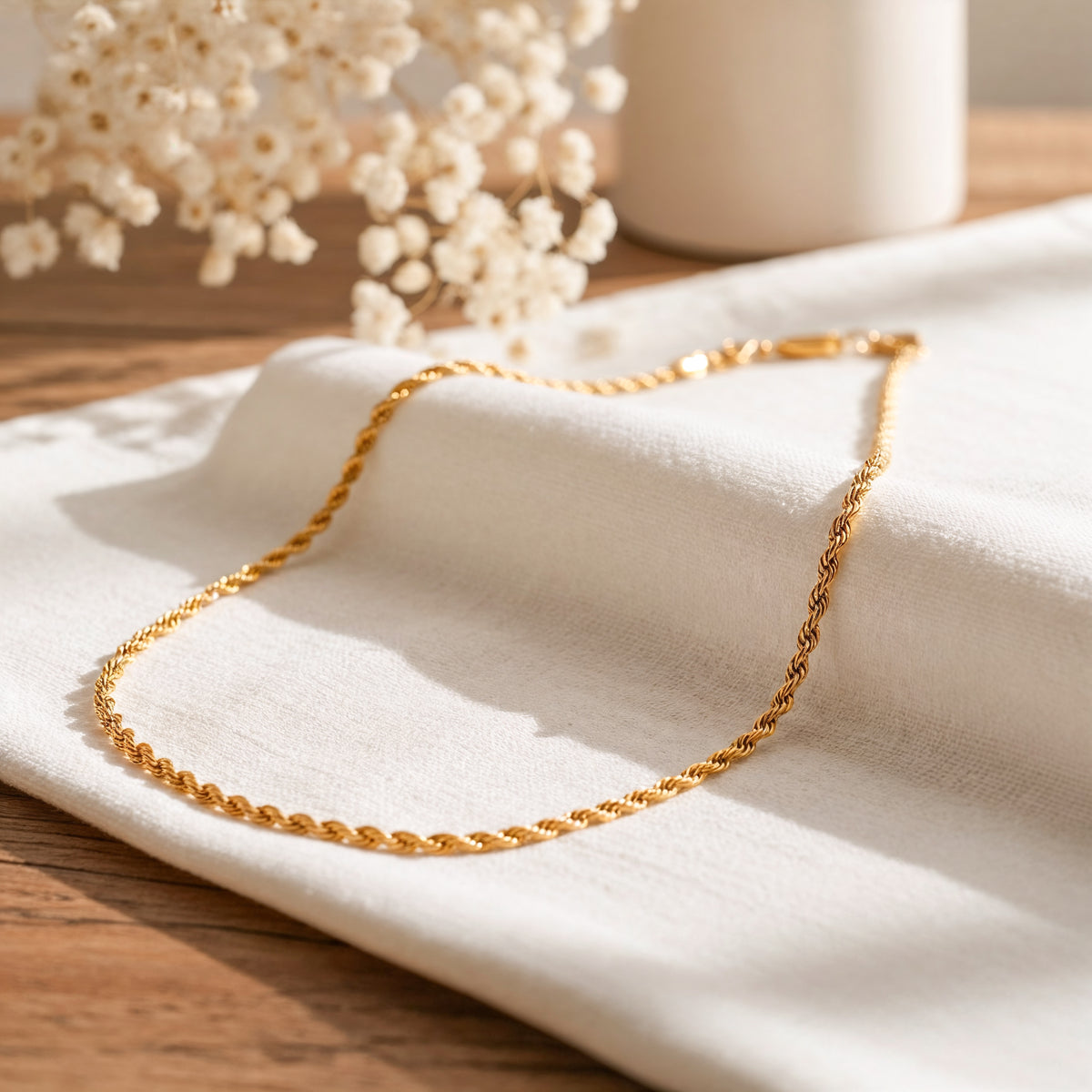 Goldene Kordel-Halskette aus Edelstahl 18 K vergoldet, liegt auf weißem Stoff mit Holzhintergrund und getrockneten Blumen – Tharien Halskette | Mercy’s Jewelry