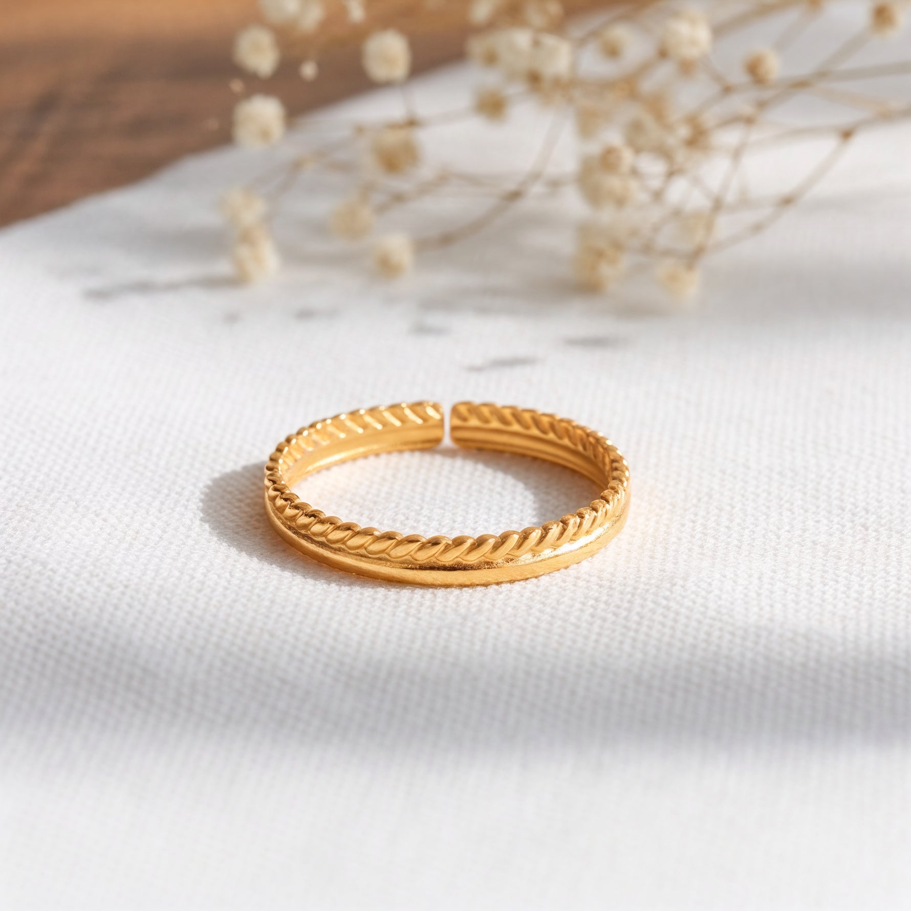 Kordel-Ring aus Edelstahl 18 K vergoldet, offen und verstellbar, mit geflochtener Struktur auf hellem Stoff mit Trockenblumen im Hintergrund – Yalith Ring | Mercy’s Jewelry
