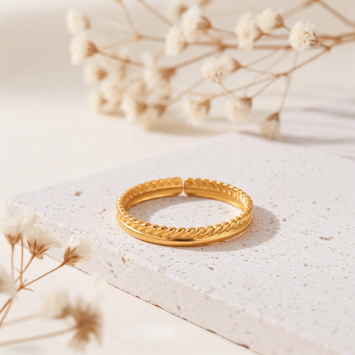 Kordel-Ring aus Edelstahl 18 K vergoldet, offen und verstellbar, mit geflochtener Struktur auf heller Steinplattform mit Trockenblumen im Hintergrund – Yalith Ring | Mercy’s Jewelry