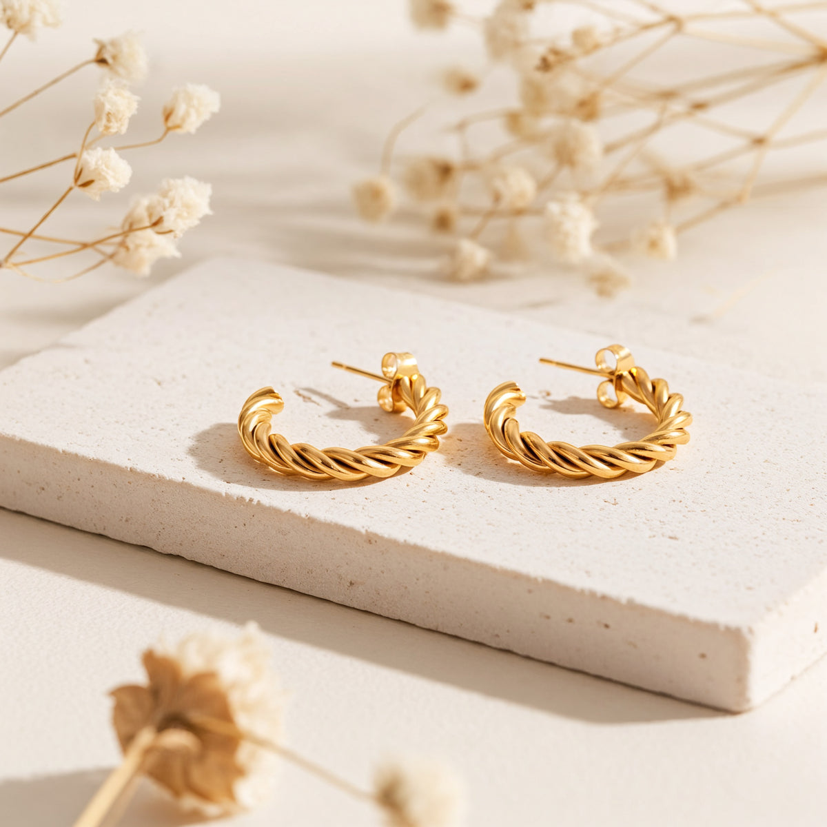 Gedrehte Kreolen aus Edelstahl 18 K vergoldet, liegen auf heller Steinplatte mit Trockenblumen im Hintergrund im warmen Sonnenlicht – Rilani Kreolen | Mercy’s Jewelry