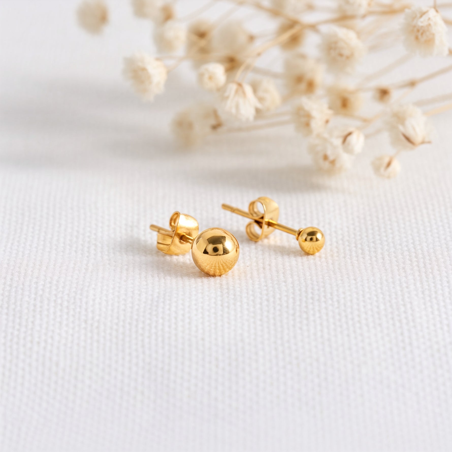Kugel-Ohrstecker aus Edelstahl 18 K vergoldet, liegen auf hellem Stoff mit Trockenblumen im Hintergrund – Mino Studs | Mercy’s Jewelry