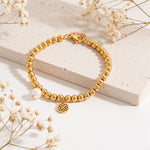 Kugelarmband aus Edelstahl 18 K vergoldet mit Ornament-Anhänger und Zierperle, stilvoll auf hellem Stein mit Trockenblumen – Solina Armband | Mercy’s Jewelry