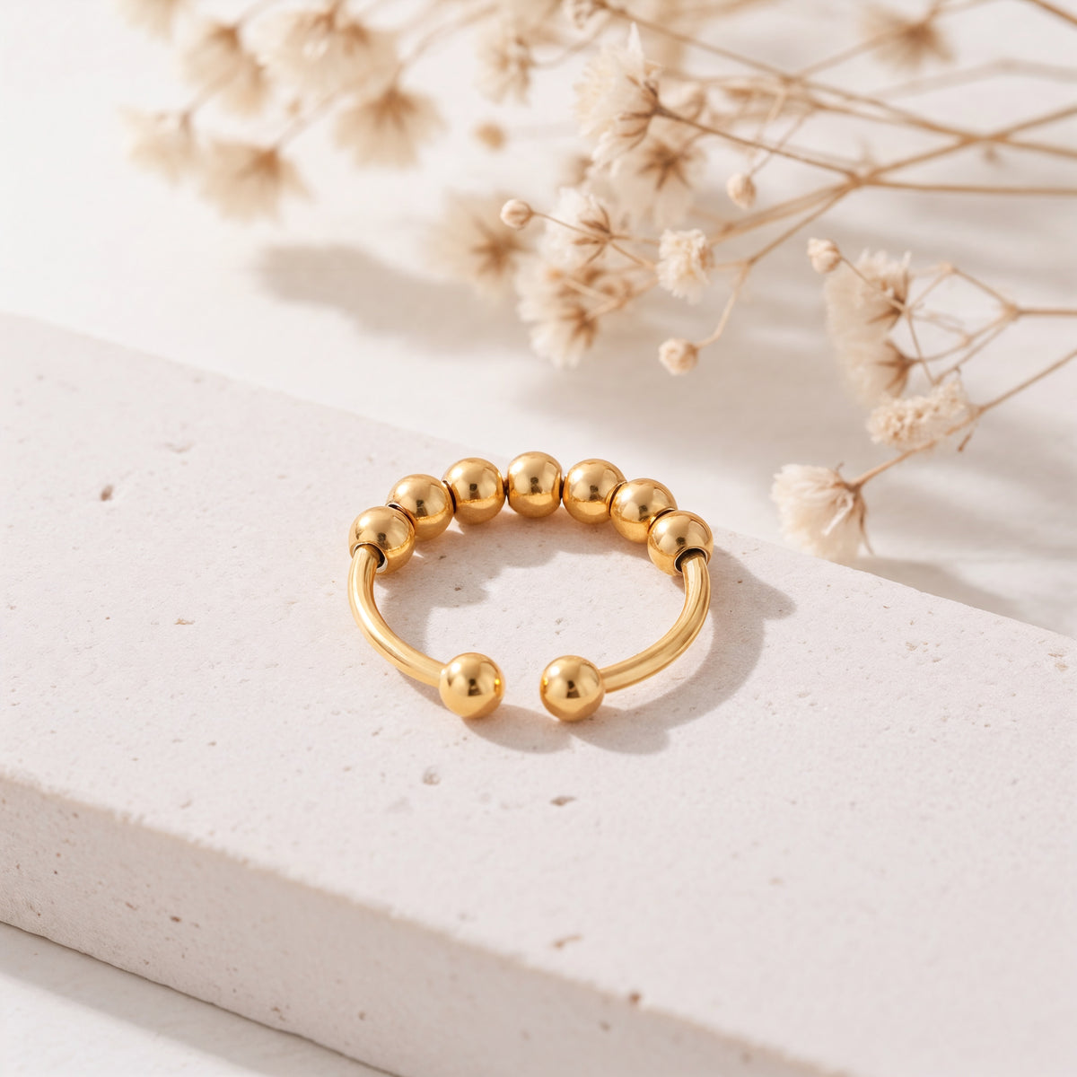 Kugelring aus Edelstahl 18 K vergoldet, offenes Design mit polierten Rundperlen auf heller Steinplattform mit Trockenblumen im Hintergrund – Yariel Ring | Mercy’s Jewelry