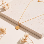 Goldene Medaillon-Halskette aus Edelstahl 14 K vergoldet, liegt auf heller Steinplatte mit getrockneten Blumen – Caleni Halskette | Mercy’s Jewelry