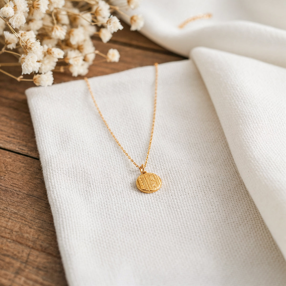 Goldene Medaillon-Halskette aus Edelstahl 14 K vergoldet, liegt auf weißem Stoff mit Holzhintergrund und getrockneten Blumen – Caleni Halskette | Mercy’s Jewelry