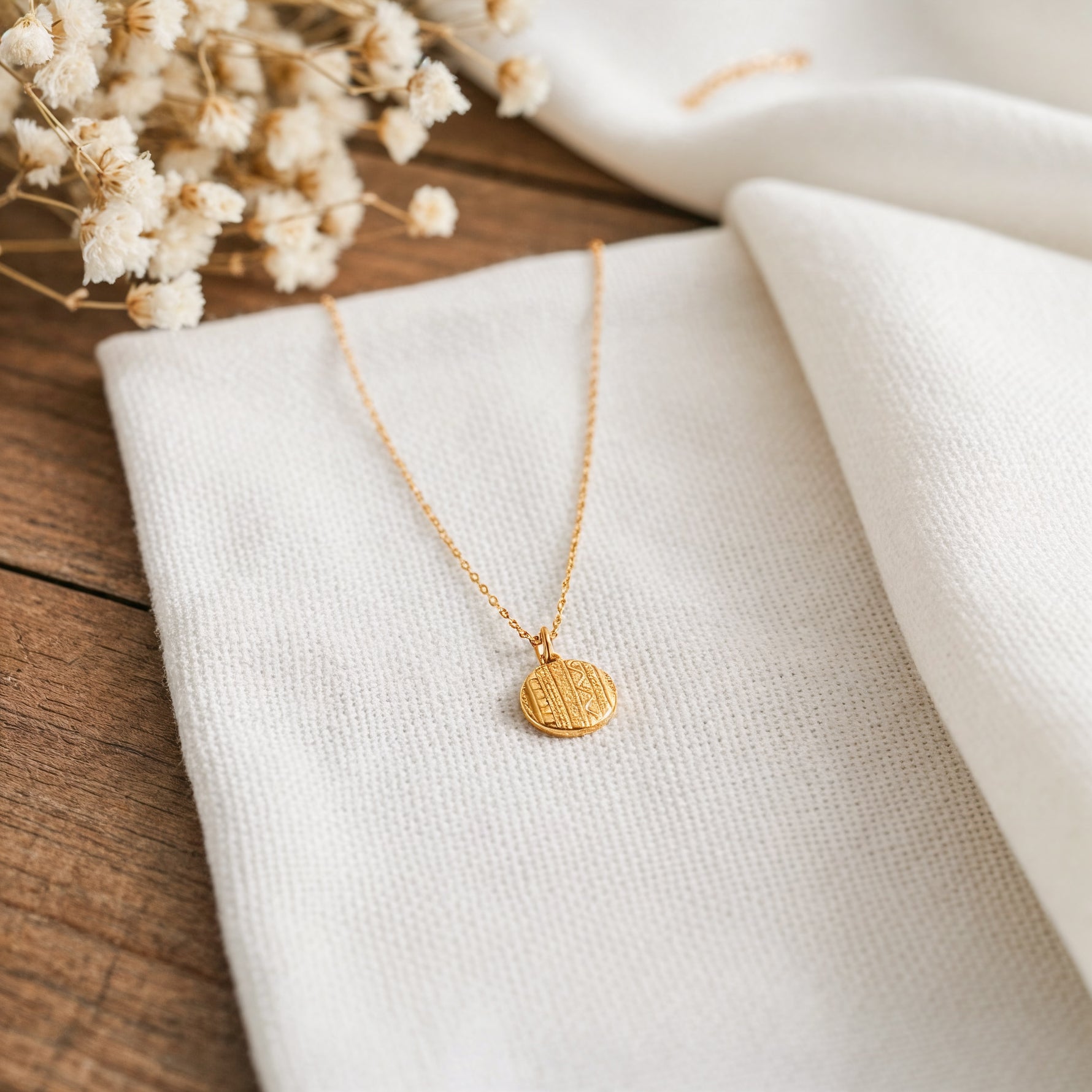Goldene Medaillon-Halskette aus Edelstahl 14 K vergoldet, liegt auf weißem Stoff mit Holzhintergrund und getrockneten Blumen – Caleni Halskette | Mercy’s Jewelry