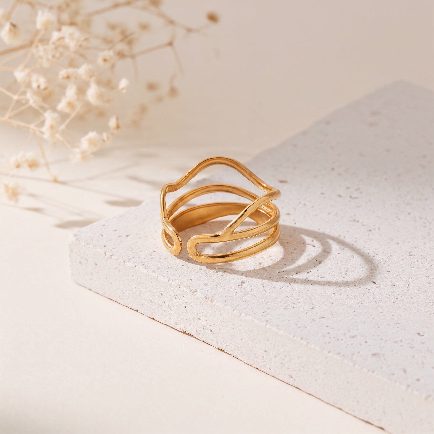 Mehrreihiger Ring aus Edelstahl 18 K vergoldet, offen und verstellbar, mit geschwungenem, modernen Design auf heller Steinplattform und Trockenblumen im Hintergrund – Tavinea Ring | Mercy’s Jewelry