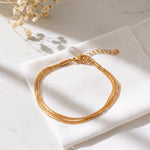 Mehrreihiges Armband aus Edelstahl 18 K vergoldet mit feinen, geflochtenen Strängen, elegant auf weißem Stoff mit sanften Schatten – Vianara Armband | Mercy’s Jewelry