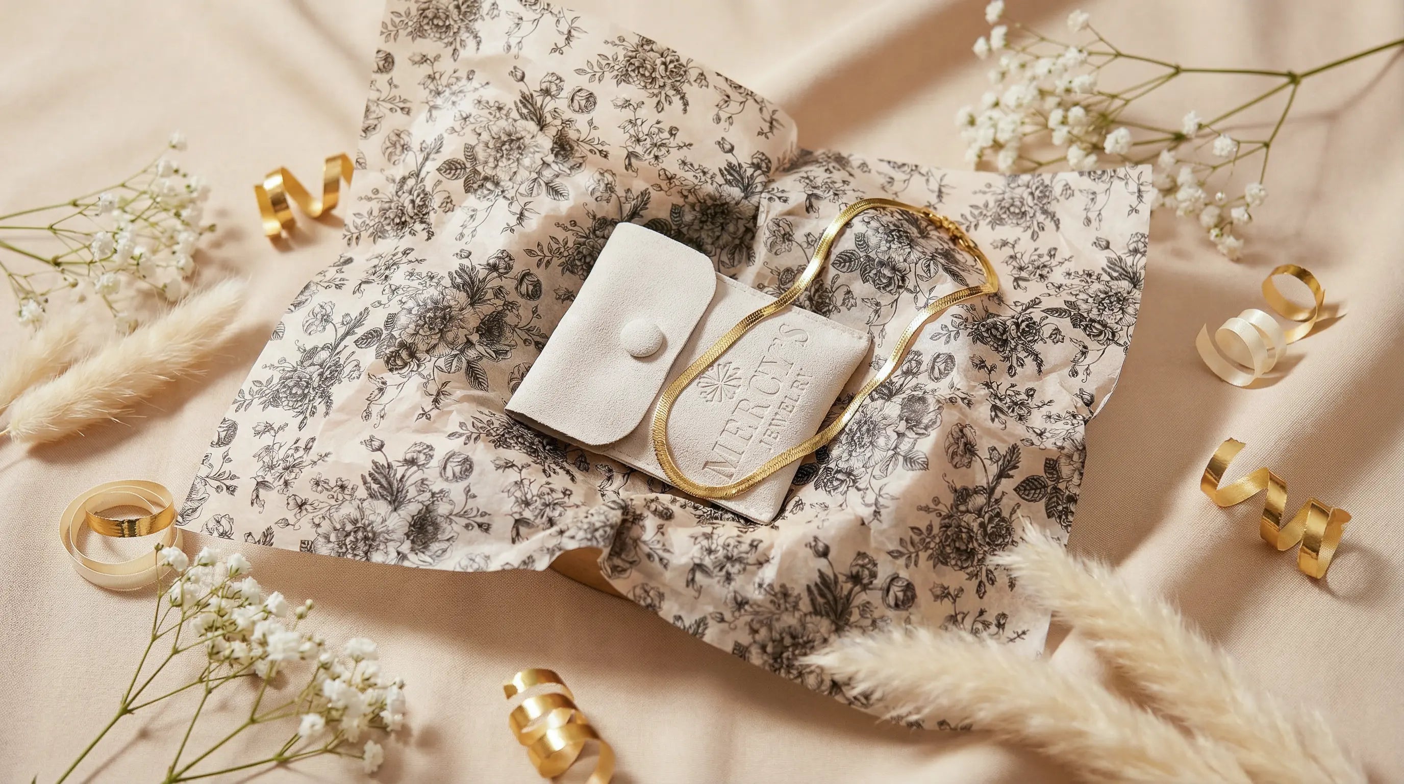 Geschenkidee bis 50 Euro von Mercy’s Jewelry: goldene Halskette auf floralem Seidenpapier, kombiniert mit hellem Schmuckbeutel, Trockenblumen und goldenen Geschenkband-Details in einer hochwertigen Flatlay-Szene.