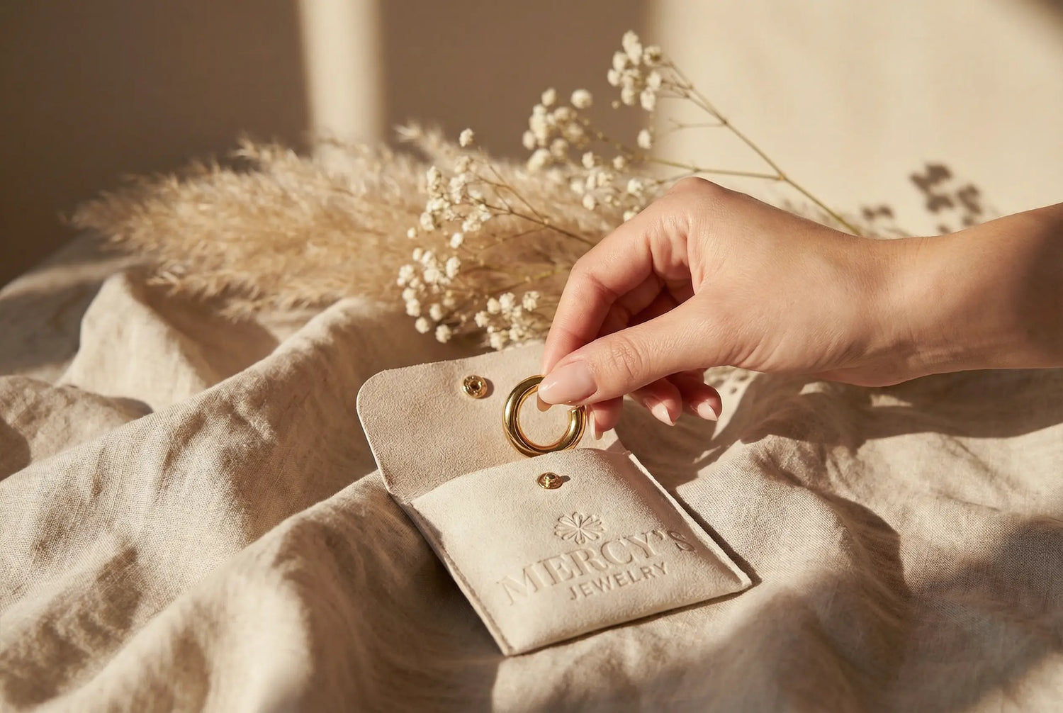 Hand legt einen goldenen Hoop-Ohrring in ein beiges Mercy’s Jewelry Pouch, dekoriert mit Pampasgras und Baby’s Breath im warmen Sonnenlicht.