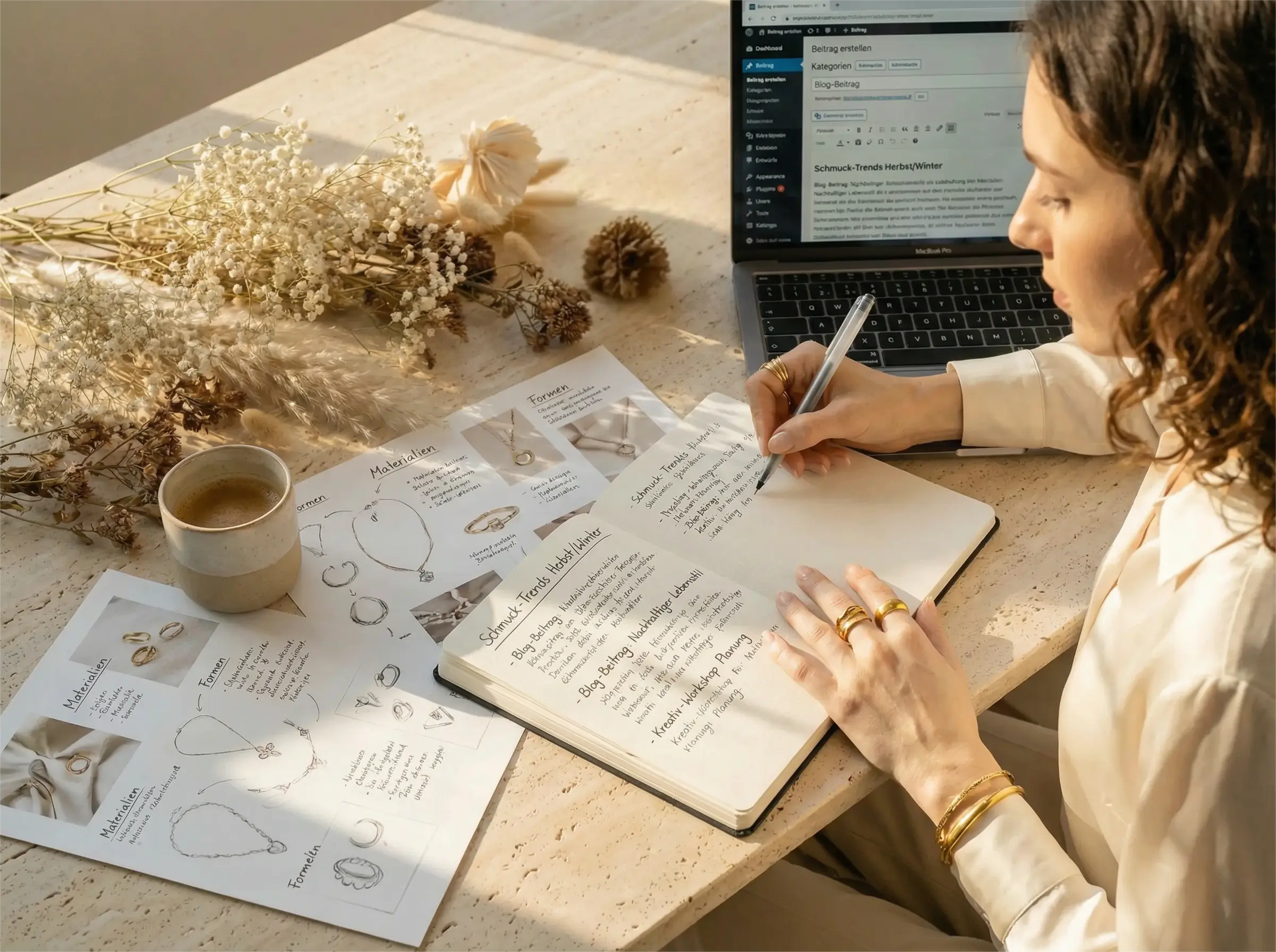 Eine Frau schreibt konzentriert in ein Notizbuch für das Mercy Journal, umgeben von Schmuckskizzen und wasserfestem Edelstahlschmuck auf einem hellen Travertin-Tisch.