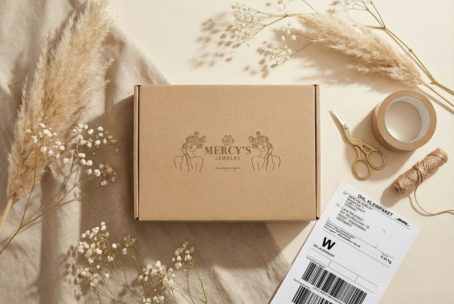 Versandkarton von Mercy’s Jewelry in warmem Flatlay-Setting mit Pampasgras, Schleierkraut und DHL-Versandlabel daneben.