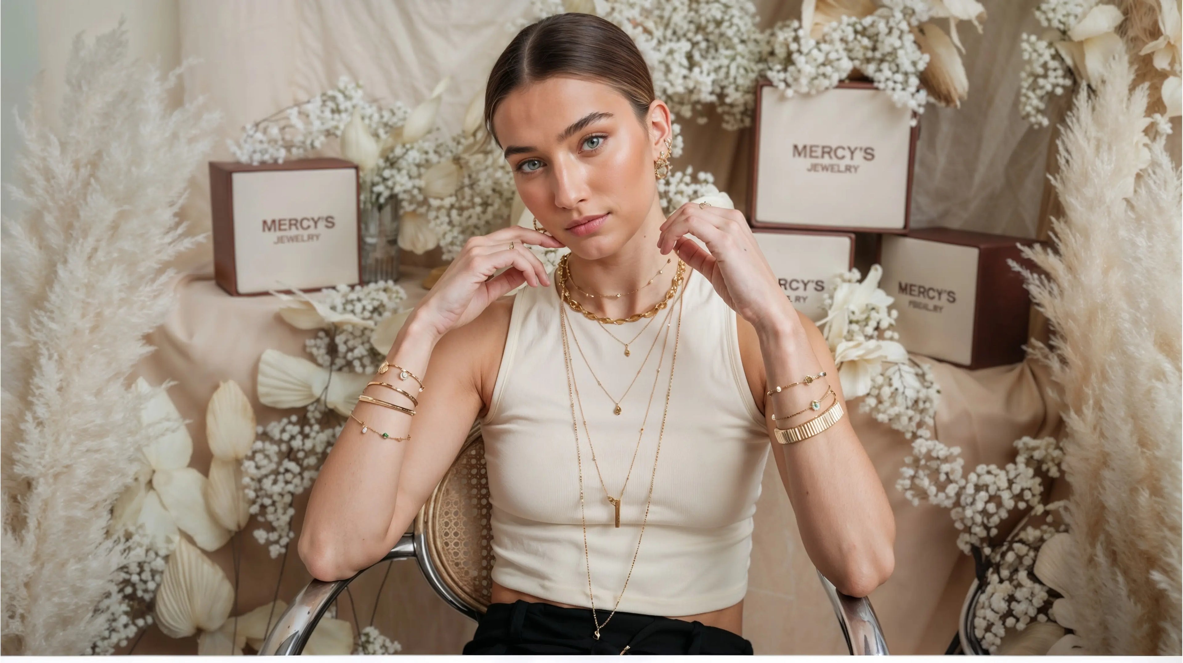 Model trägt Mercy’s Jewelry Bestseller mit goldenen Halsketten im Layering-Look, Armbändern, Creolen-Ohrringen und Ringen, stilvoll präsentiert vor dekorativen Blumen und Schmuckboxen