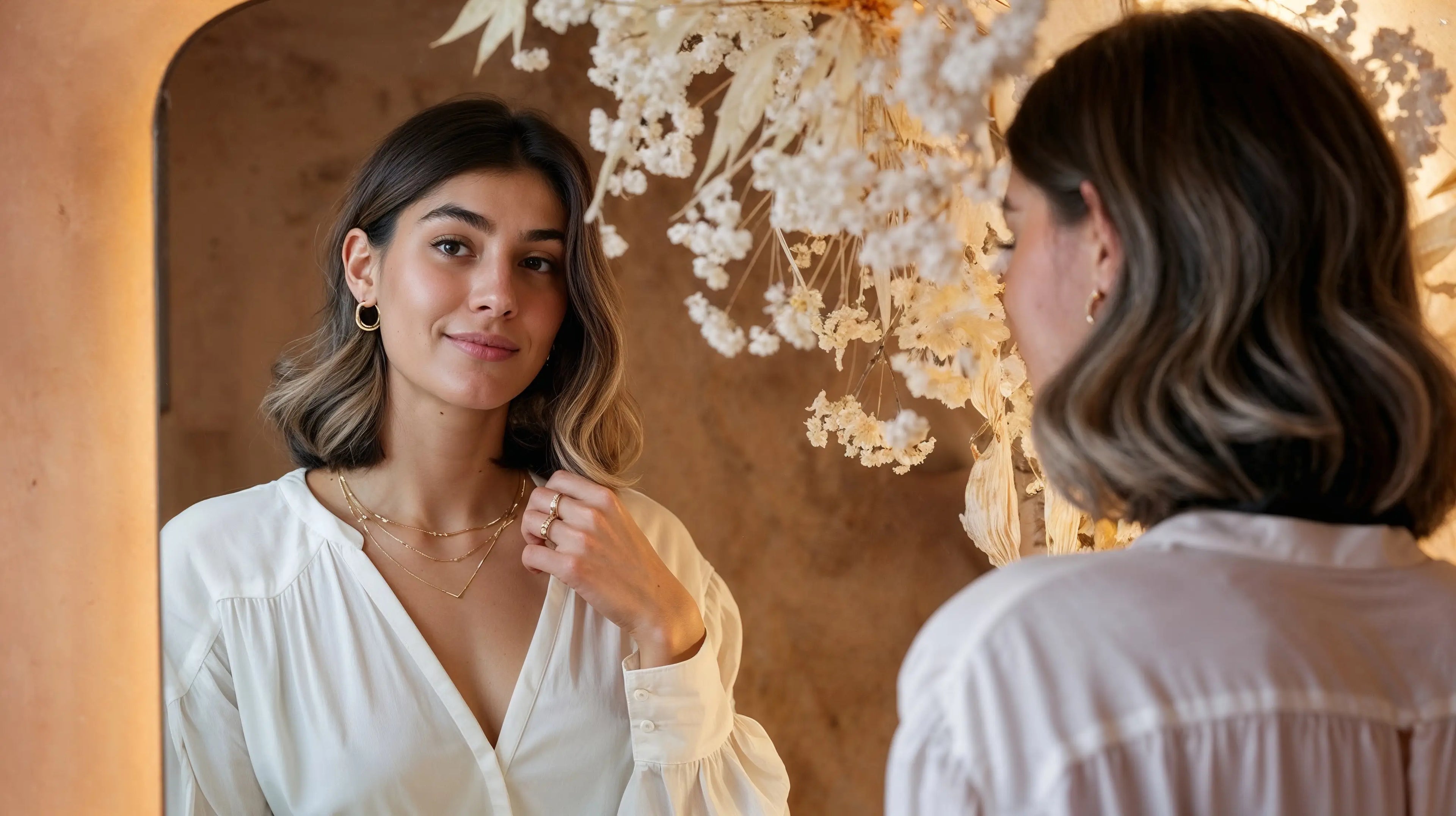Model trägt Mercy’s Jewelry Essentials mit goldenen Halsketten, Creolen-Ohrringen und Ringen und betrachtet sich im Spiegel, umgeben von dekorativen Trockenblumen