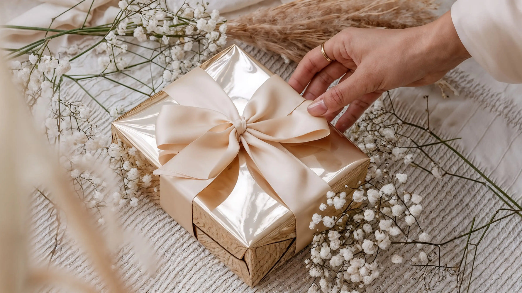 Hand legt eine goldene Geschenkbox mit cremefarbener Schleife von Mercy’s Jewelry zwischen weißen Blüten auf Stoffuntergrund