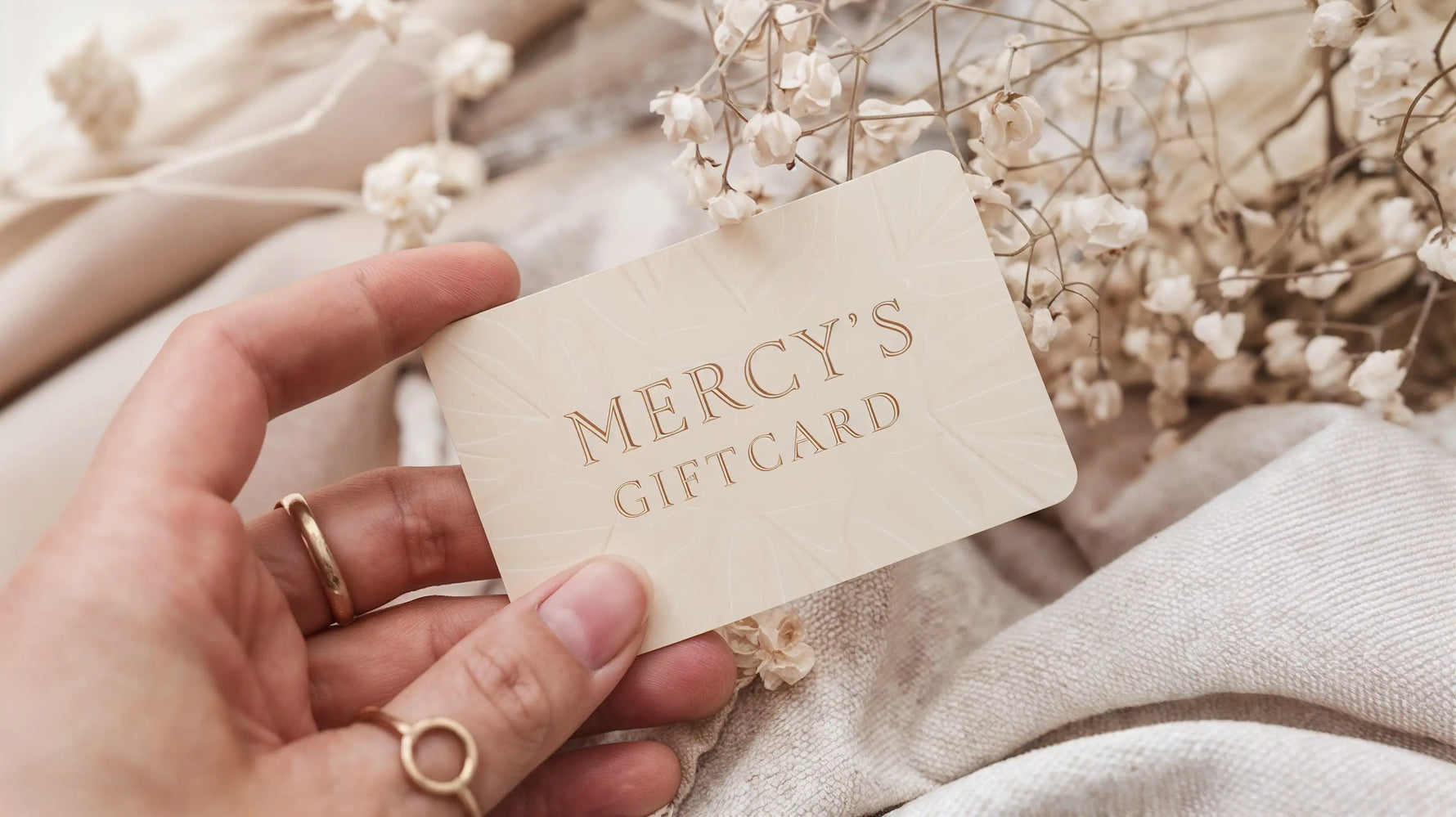 Hand hält eine Mercy’s Jewelry Geschenkkarte in Cremefarbe vor Stoff und weißen Blüten