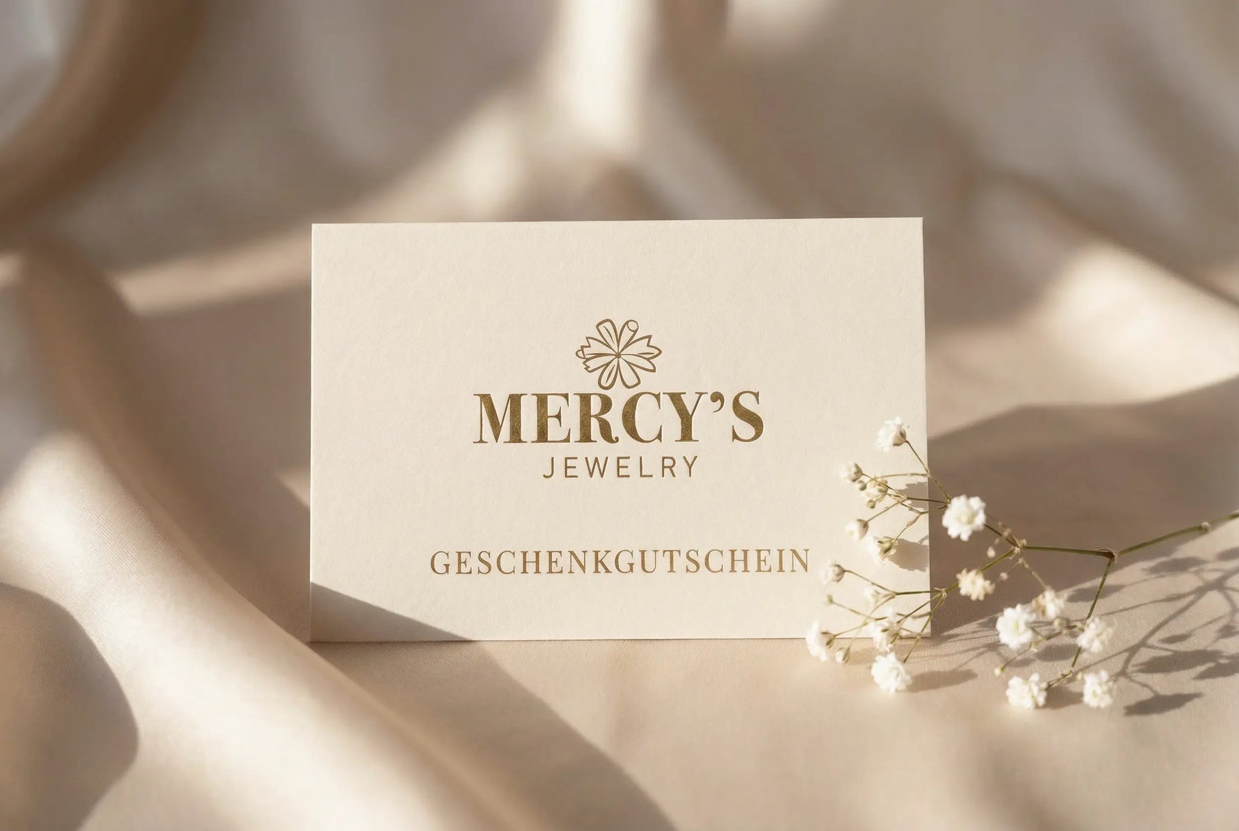 Mercy’s Jewelry Geschenkgutschein auf cremefarbener Karte, platziert auf satiniertem Stoff mit zartem Schleierkraut
