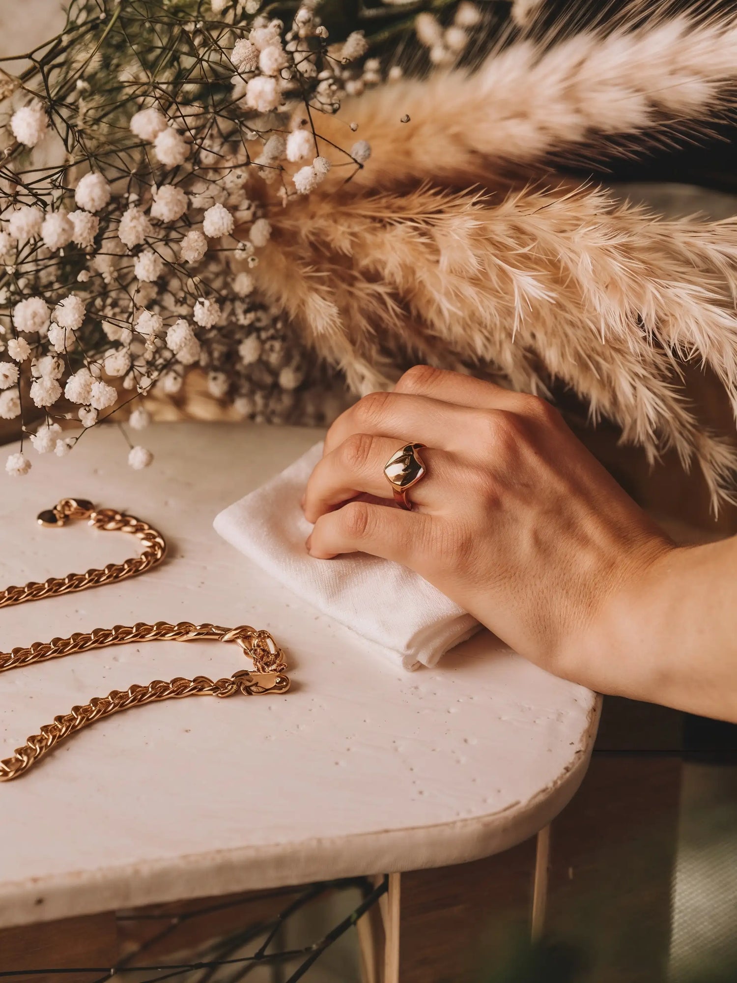 Hand mit Mercy’s Jewelry goldenem Ring poliert goldene Kette mit weißem Tuch auf Tisch, Blumendekoration im Hintergrund